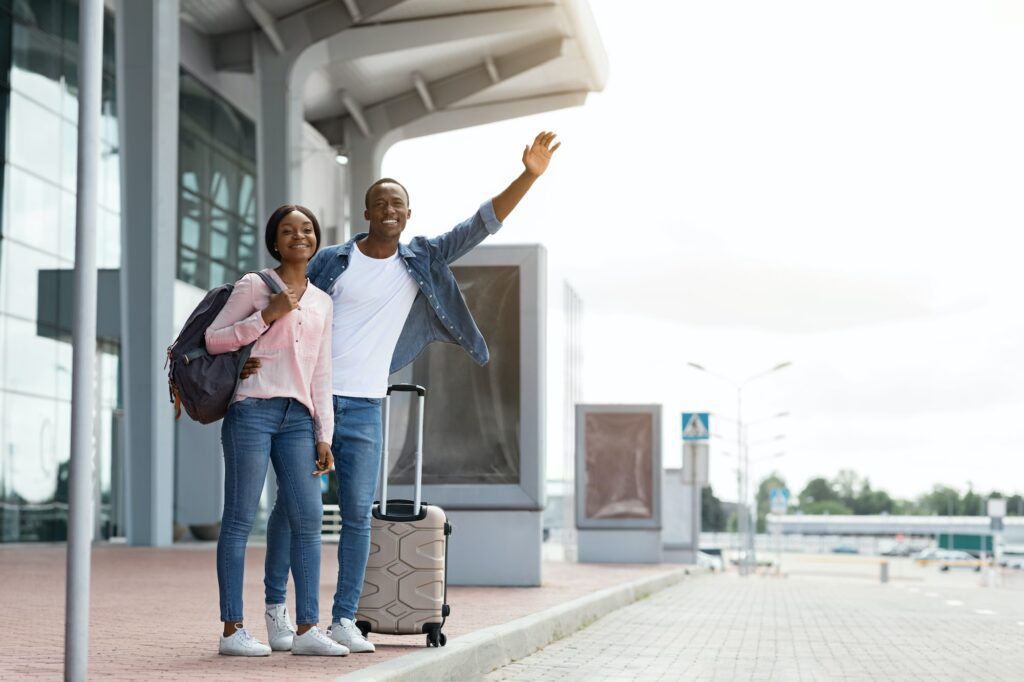 Happy Young Black Couple Standing With Suitcases Near Airport Terminal, Catching Taxi