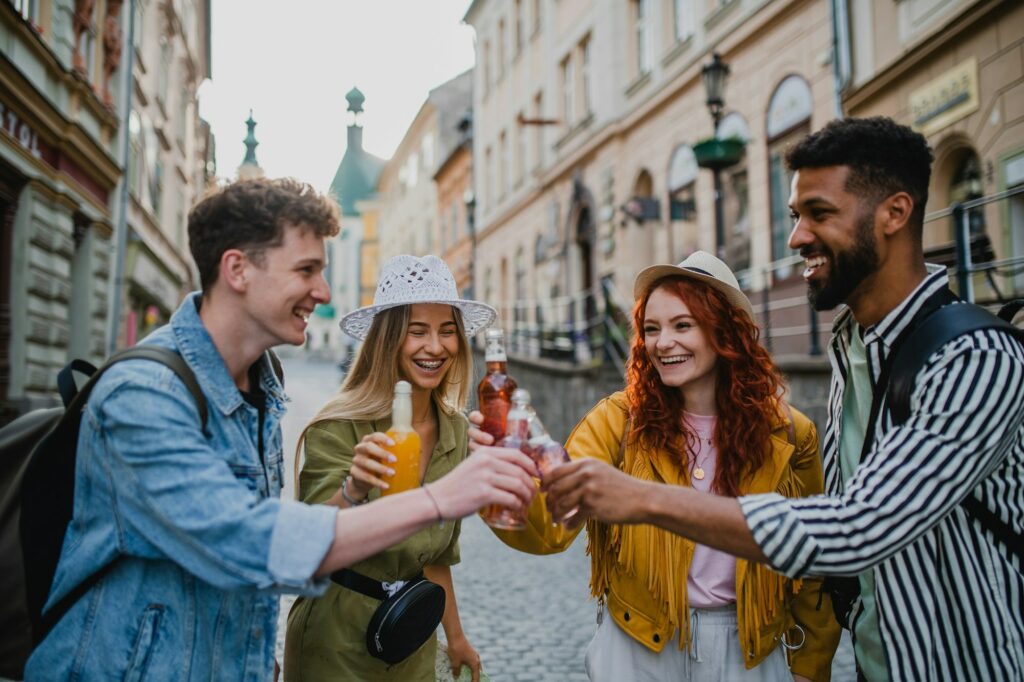 Front view of group of happy young people with drinks outdoors on street on town trip, laughing